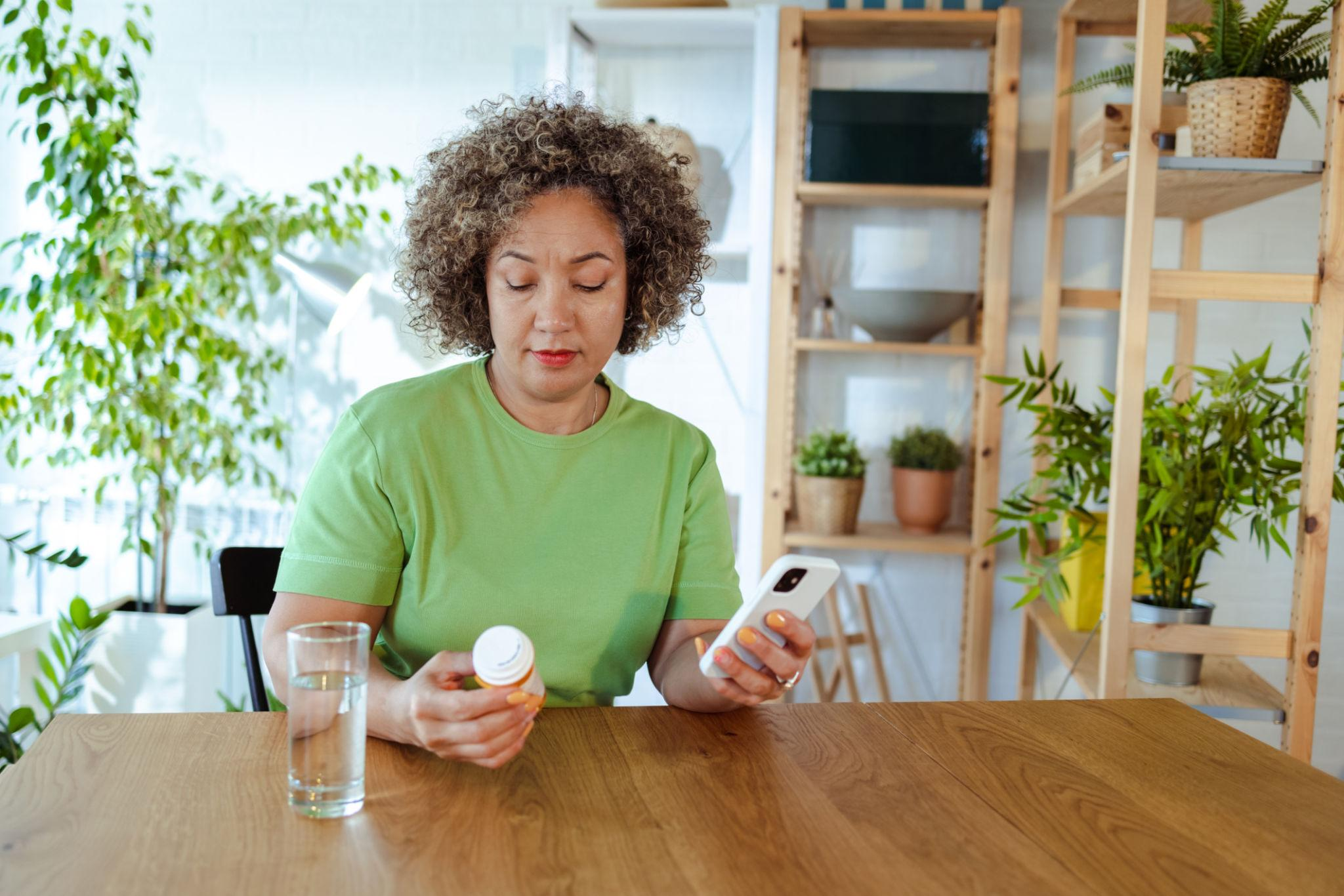 A woman at home checking a medication bottle while using her phone
